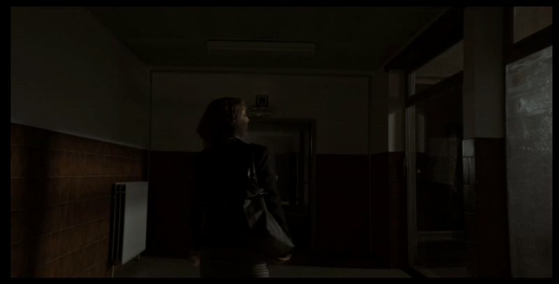 A blonde short haired woman is walking her university halls at night, a car illuminates the passage and a man can be seen following behind her.